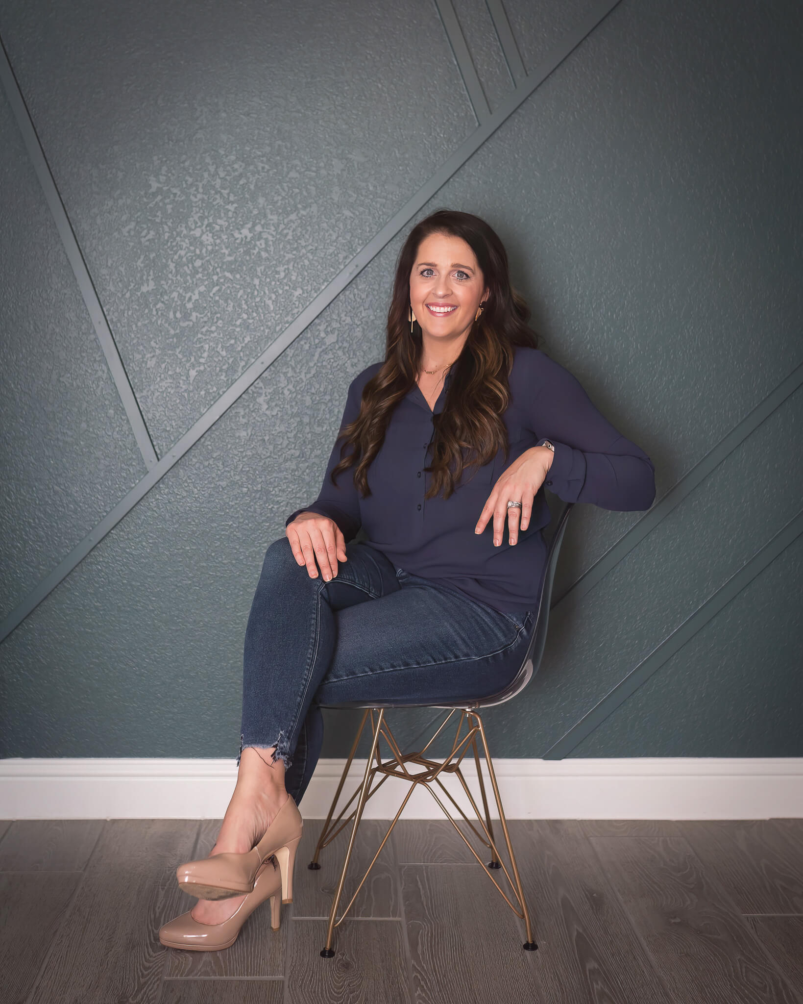 business owner in clear chair in navy blue top with jeans and beige heels. dark teal feature wall as her background