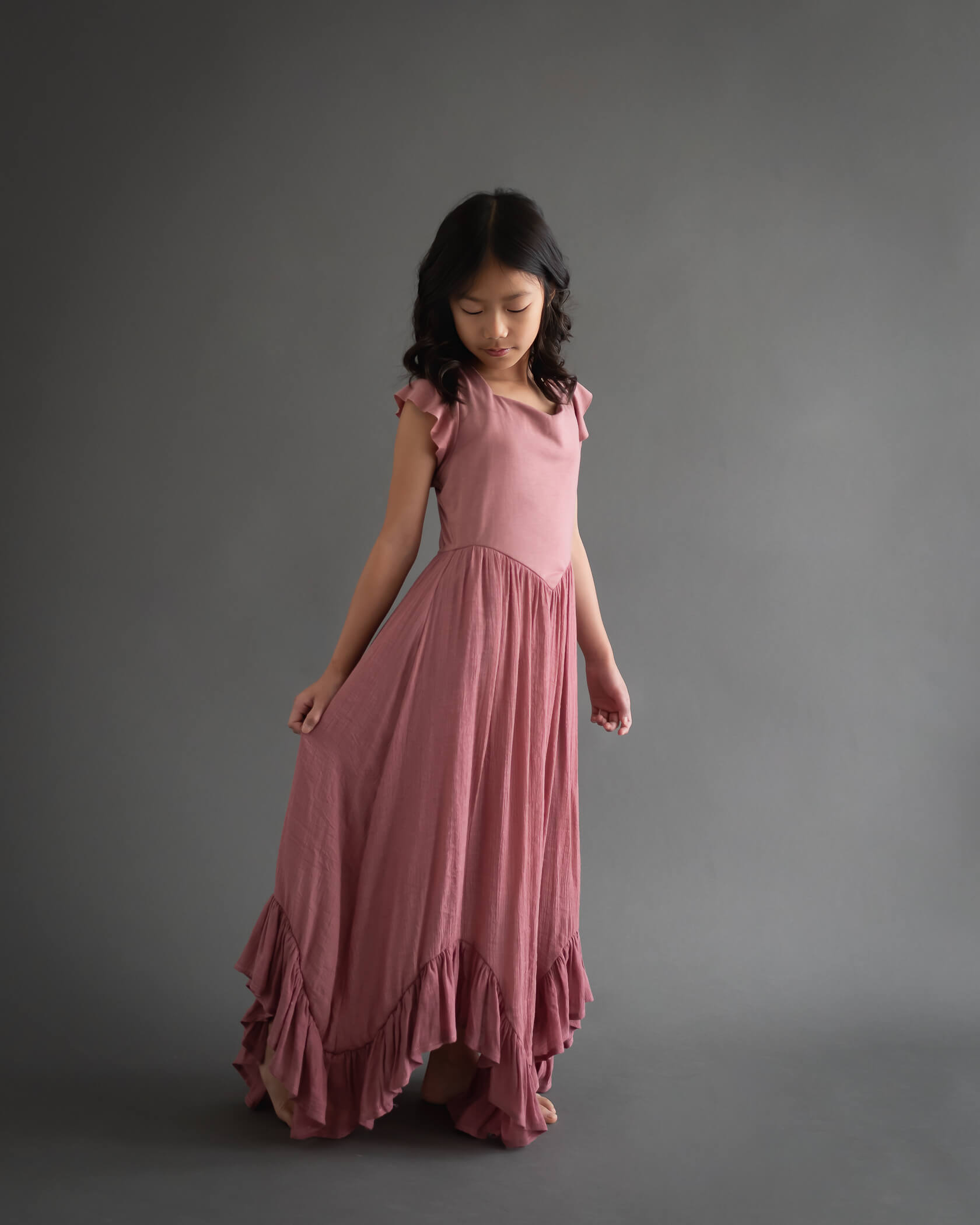 10 year old girl in studio portrait wearing a long dress holding the blush-colored dress out and looking down