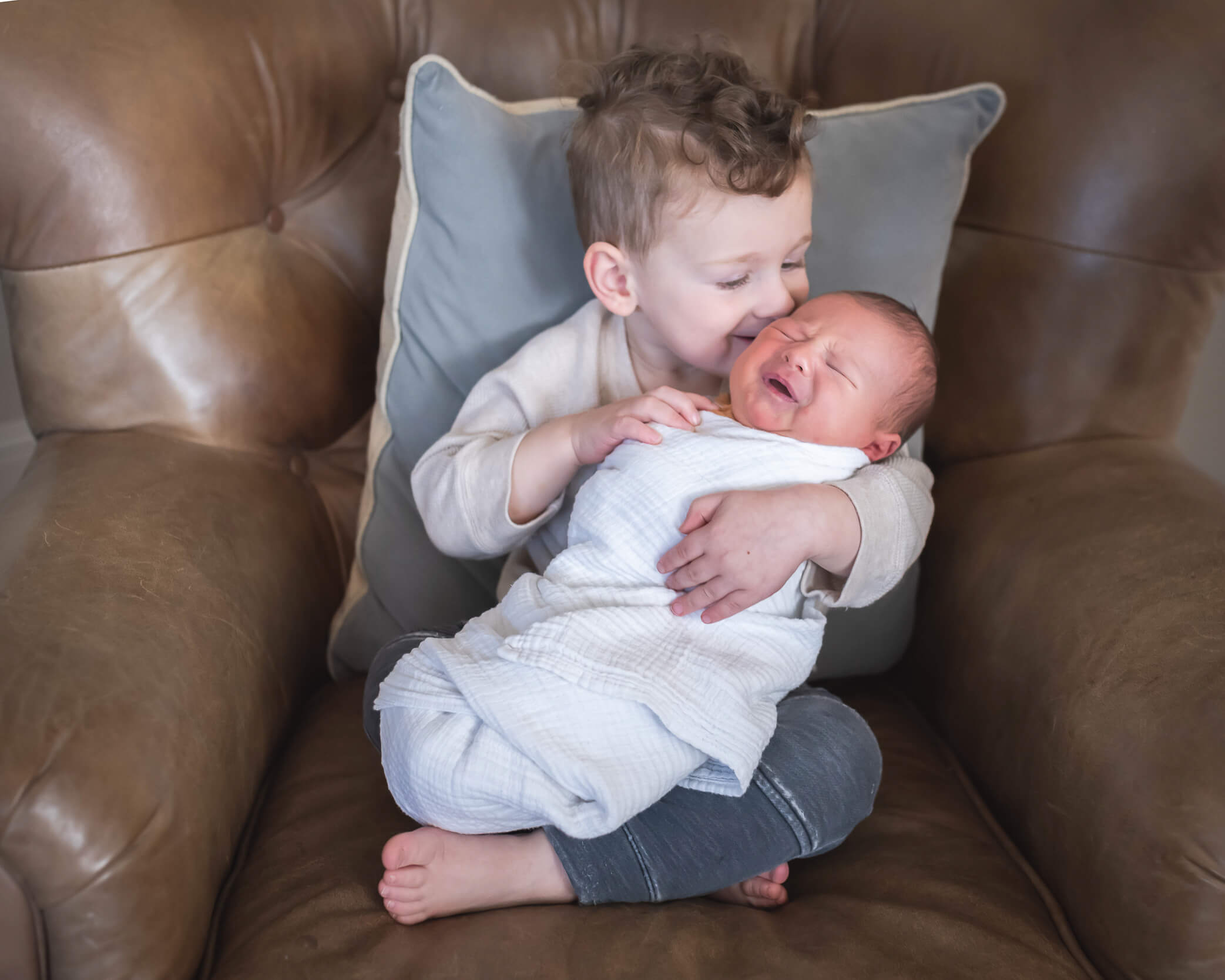 littlest brother holding and kissing his new baby sibling in a comfy leather chair