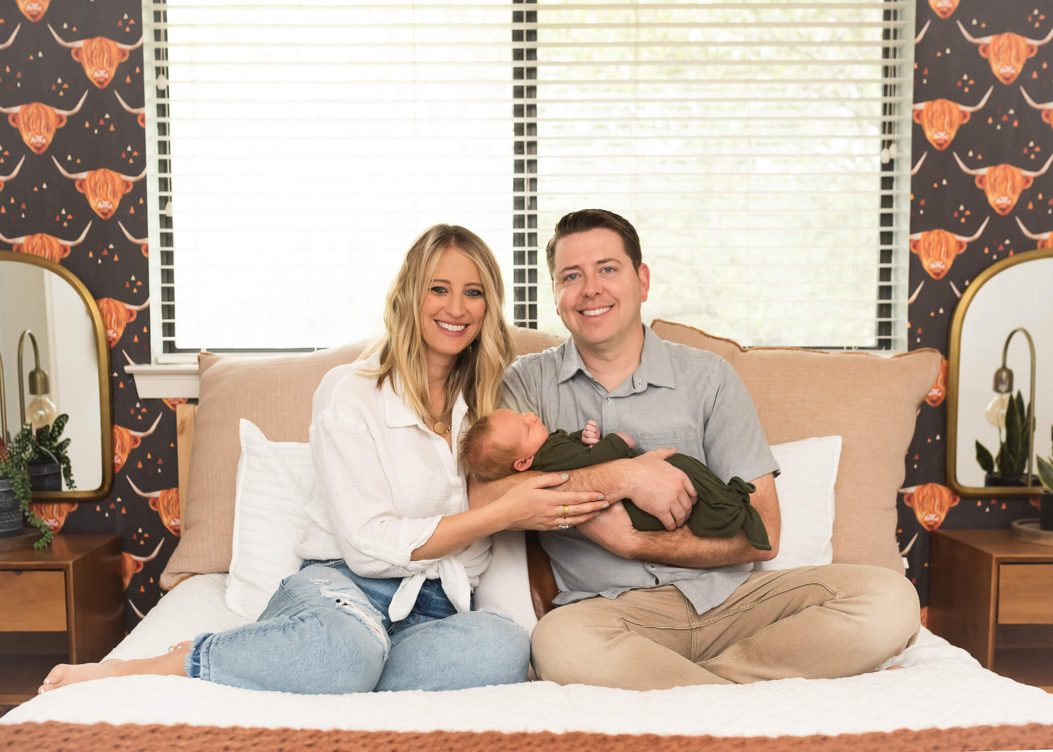 parents on bed looking at photographer smiling, so happy with new baby bow
