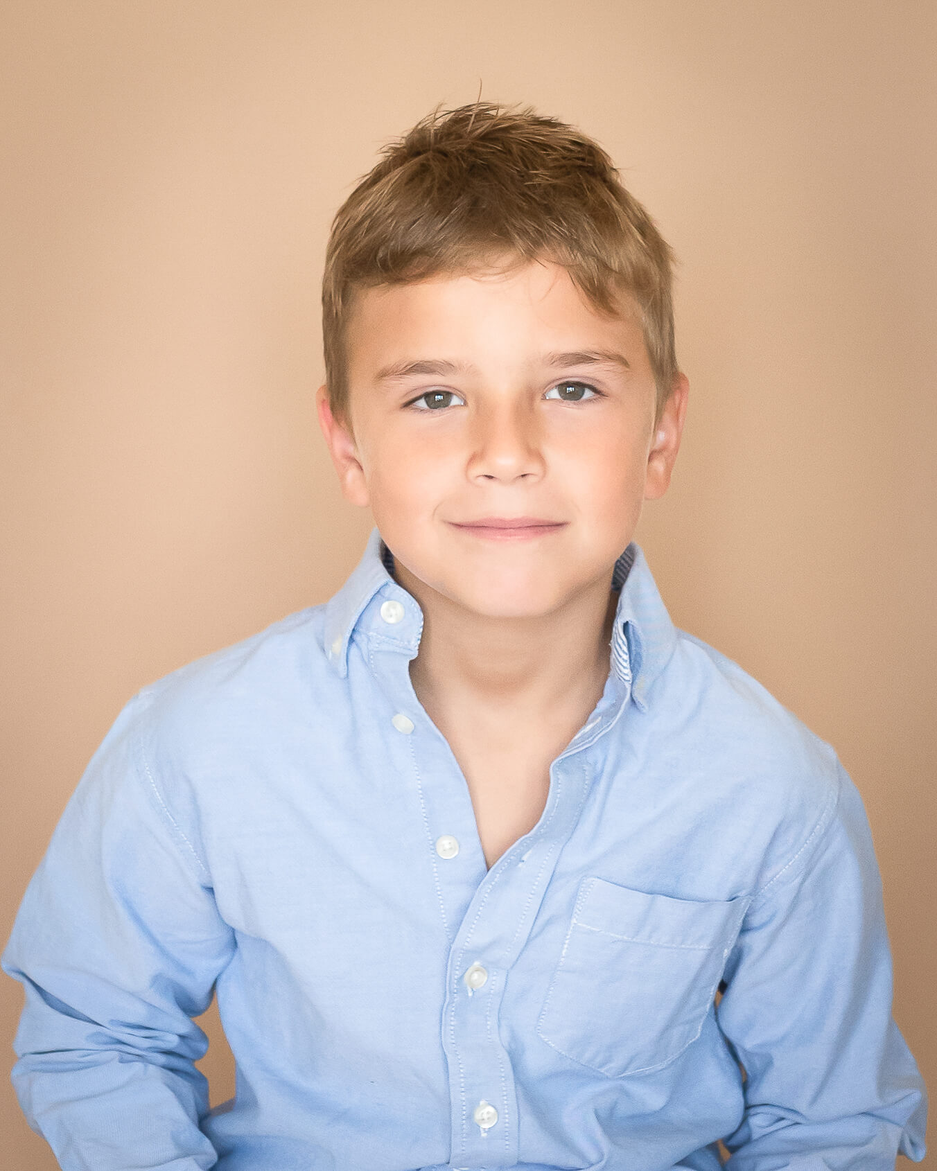 studio portrait of an 8 year old boy with a blue dress shirt on a tan background