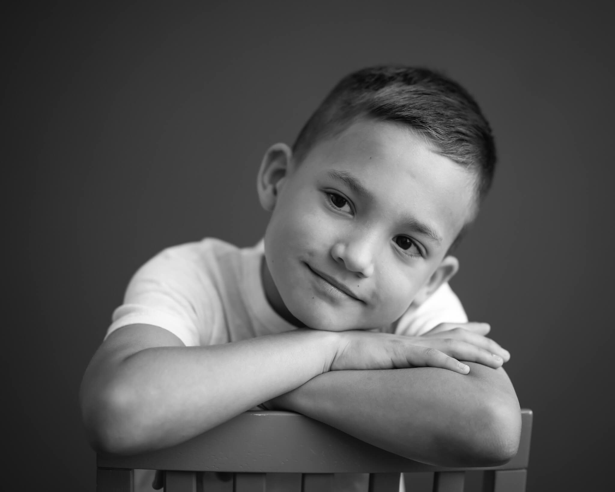 handsome 8 year old boy resting head on crossed arms over the back of chair in black and white studio portrait