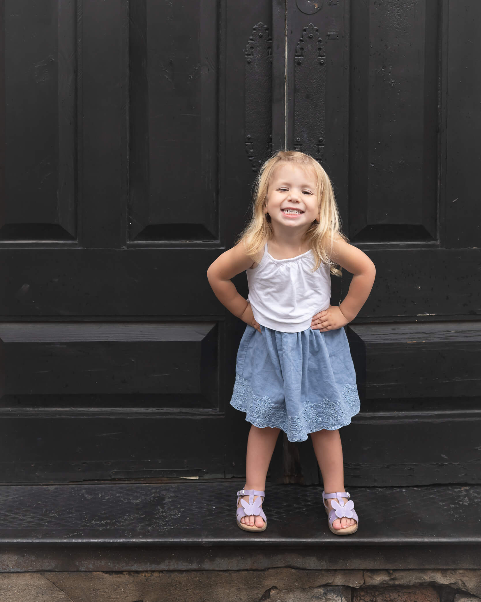 Precious little girl in white tank top and denim skirt in front of black door in Downtown McKinney captured by Allison Amores Photography