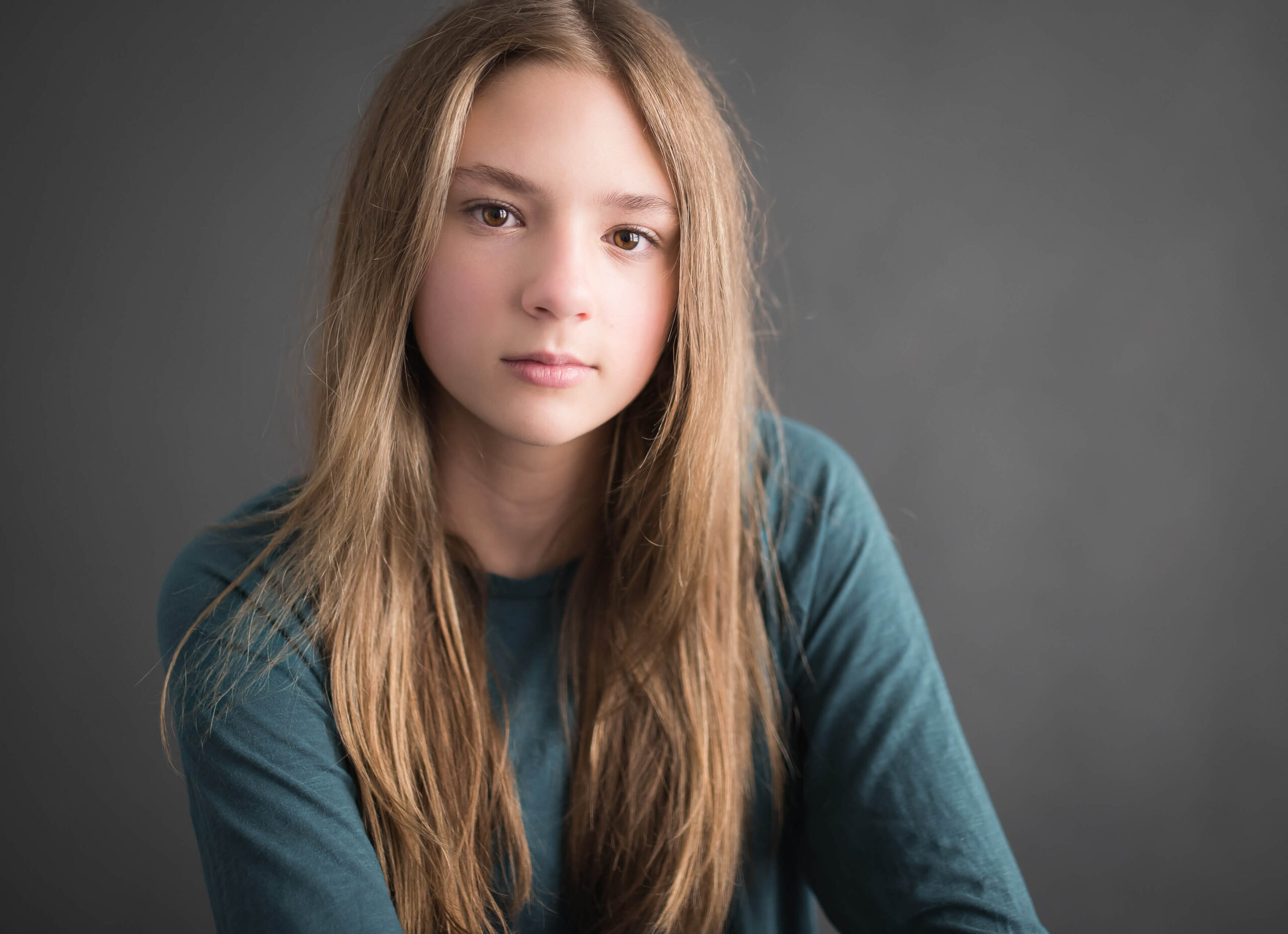 Preteen performing arts headshot of girl with long blonde hair in teal long-sleeved shirt on dark background, captured by Allison Amores Photography.